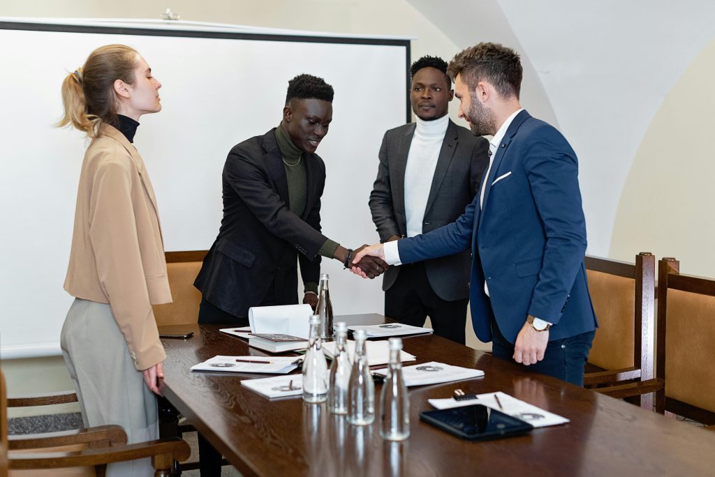 Four professionals in a modern office, two men shaking hands signifying agreement.
